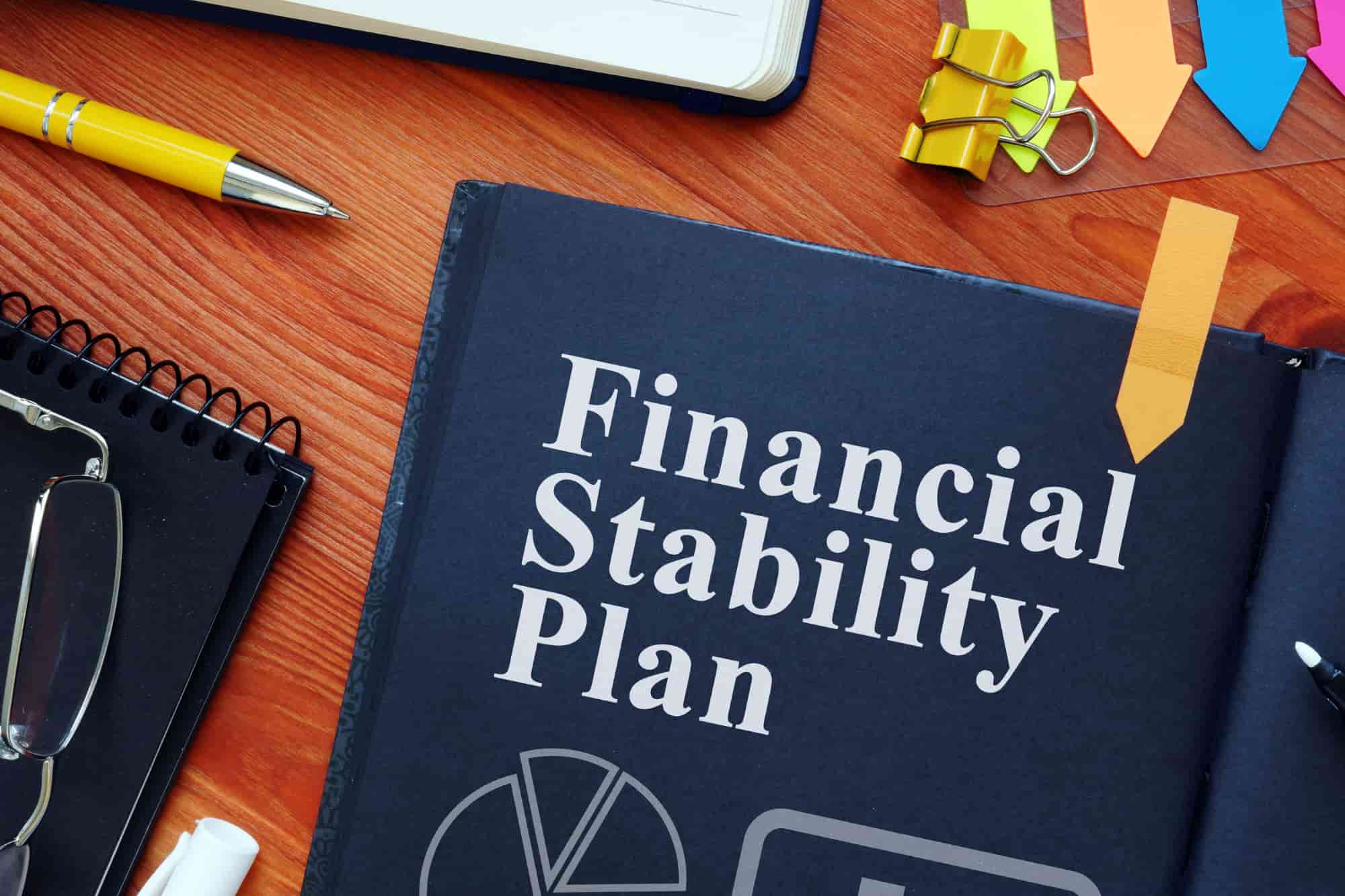 Glasses, a notebook, a pen. Post-its, a paperclip, and a book titled “Financial Stability Plan” sit on a desk