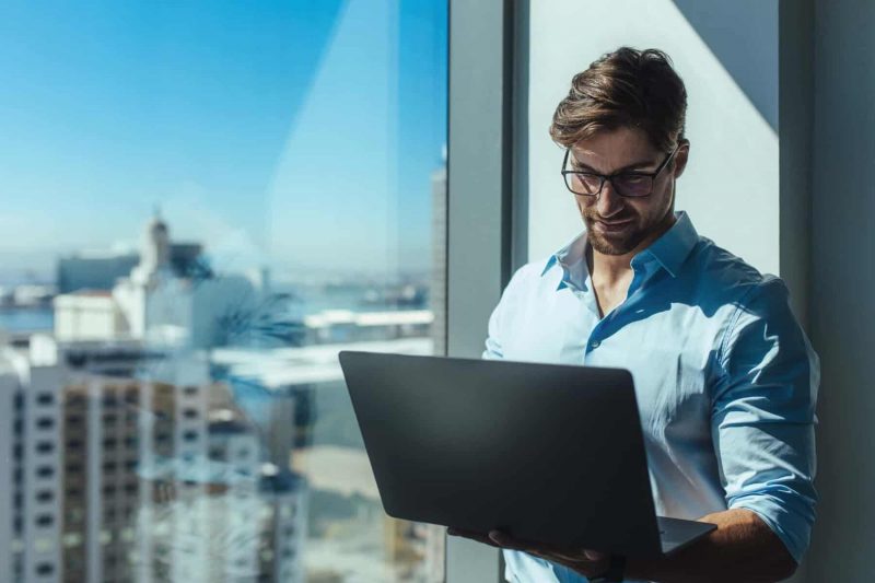 A man stands by a city window and looks at his laptop