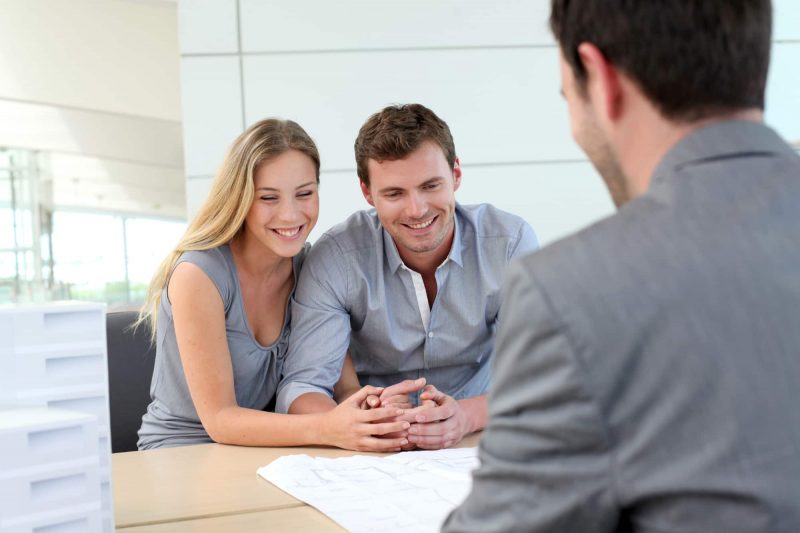 A woman and man embrace and smile at documents, the back of a man in a suit appears in the foreground