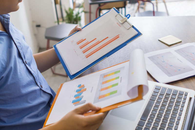 A person comparing financial charts and graphs on two clipboards, with a laptop and more papers visible on the table.