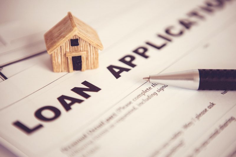 A miniature wooden house sits on a loan application form, with a pen resting on the document.