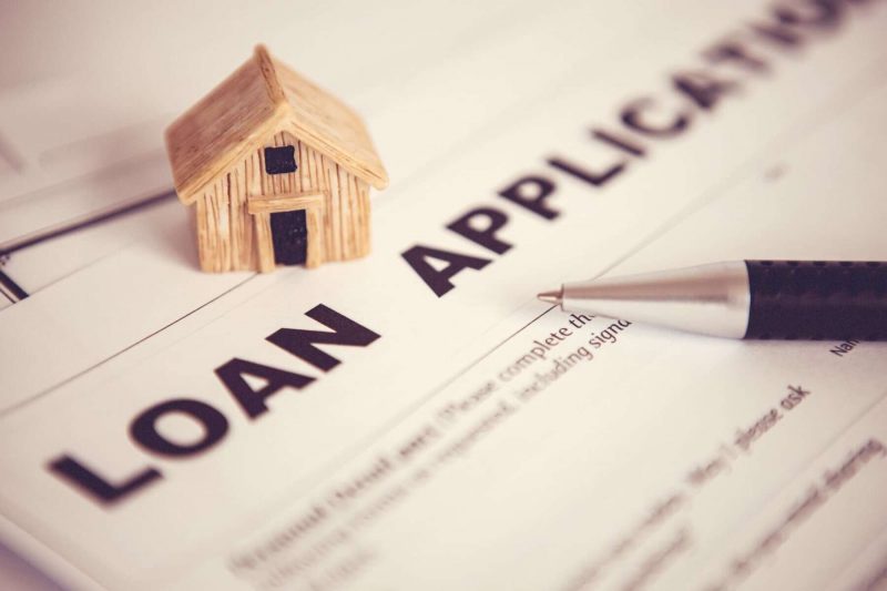 A miniature wooden house sits on a loan application form, with a pen resting on the document.